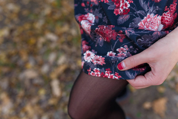 Female hand with red manicure holding dress or skirt with red flowers on blurry autumn background. Nature, health and beauty, hands  and fingernails care concept. - Photo, Image