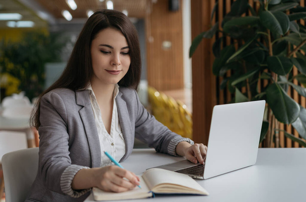 Деловая женщина, работающая в офисе. Красивая женщина-фрилансер, пользующаяся ноутбуком, печатающая, записывающая в блокнот. Портрет молодого успешного писателя
 - Фото, изображение