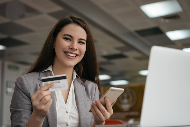 クレジットカードを持っているビジネスマン、オンラインショッピングのためのスマートフォンを使用して、オフィスに座っています。フリーランスは支払いを受け、残高を確認します。笑顔の女性は食べ物を注文し、ウェブサイト上でチケットを予約する - 写真・画像