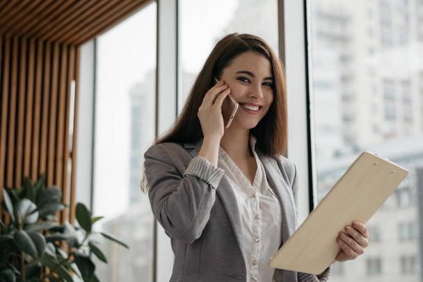 Portrait of attractive manager holding smartphone, communication, looking at camera, smiling. Confident business woman talking on mobile phone, standing in modern office - Valokuva, kuva