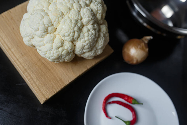 Cauliflower on chopping board, hot red pepper and onion on black kitchen table. Organic vegetarian food.  - Photo, image