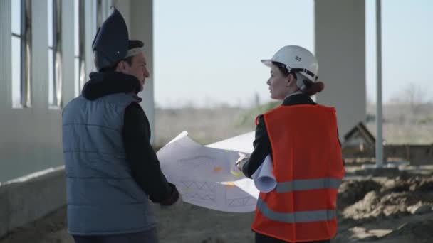 male and female industrial engineers in protective suits and helmets who are discussing construction of hangar while walking along pillar of new building background of welding sparks - Footage, Video
