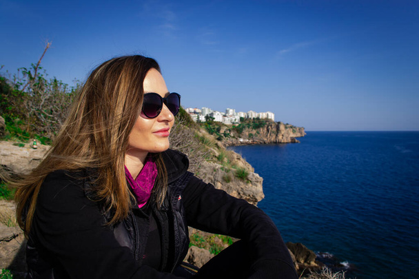 Belle jeune fille heureuse avec des lunettes de soleil assis sur le rocher relaxant et regardant vers la mer bleue
 - Photo, image