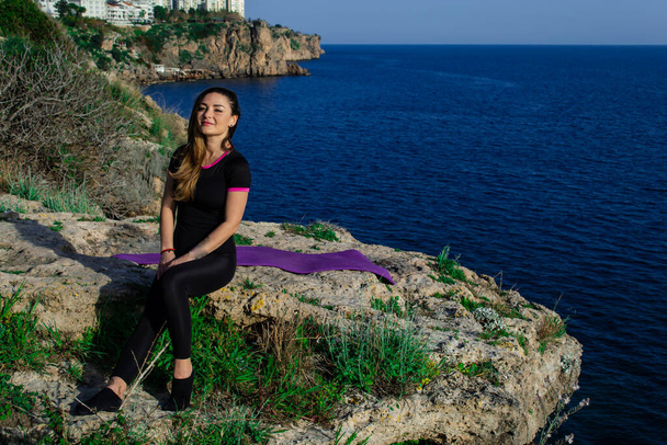 Beautiful young happy girl smiling sitting on rock near blue sea on a sunny day - Photo, Image