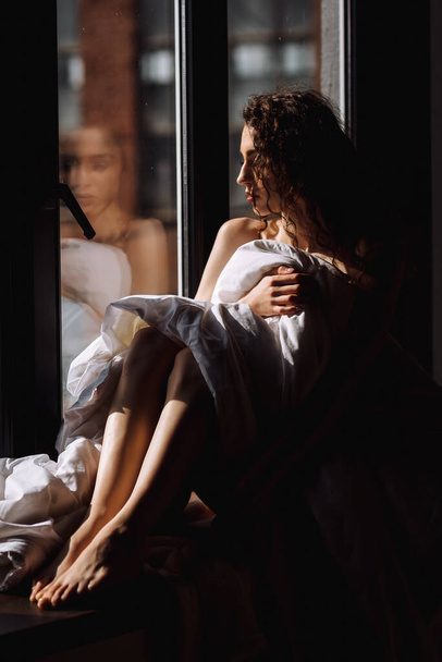 göndör, meztelen, takaróba csomagolt lány ül az ablakpárkányon. - Fotó, kép