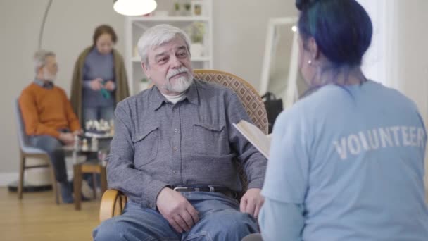 Portrait of smiling old Caucasian man listening to volunteer reading book. Young woman spending time with elderly male retiree in nursing home. - Filmagem, Vídeo