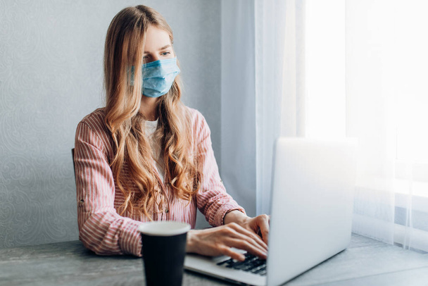 Uma jovem empresária trabalha em casa, sentada em uma mesa com um laptop, usando uma máscara protetora. O conceito de quarentena, coronavírus
 - Foto, Imagem