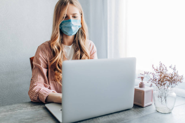 A young businesswoman works from home, sitting at a table with a laptop, wearing a protective mask. The concept of quarantine, coronavirus - 写真・画像