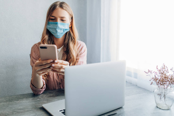A young businesswoman in a protective mask works at home during quarantine, sitting at a table with a mobile phone. Quarantine, coronavirus, - Φωτογραφία, εικόνα