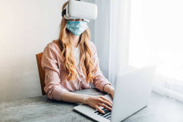 A young woman in a protective mask and virtual reality glasses, working at home during quarantine with a laptop. Quarantine, coronavirus - Foto, imagen