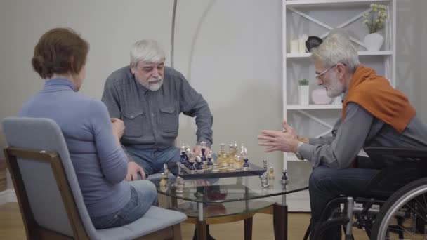 Mature grey-haired Caucasian men playing chess and talking as woman knitting in armchair. Elderly ill and disabled people spending time in nursing home. - Πλάνα, βίντεο