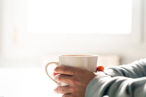 Το άτομο ζεσταίνει τα χέρια του με ένα ζεστό φλιτζάνι καφέ. Κρατώντας ένα κύπελλο. - Φωτογραφία, εικόνα