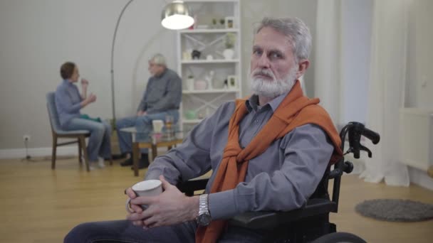 Portré egy jóképű, öreg fehér férfiról, aki tolószékben ül és a kamerába néz. Idős férfi nyugdíjas, aki más rezidensekkel tölti az estét az idősek otthonában. - Felvétel, videó