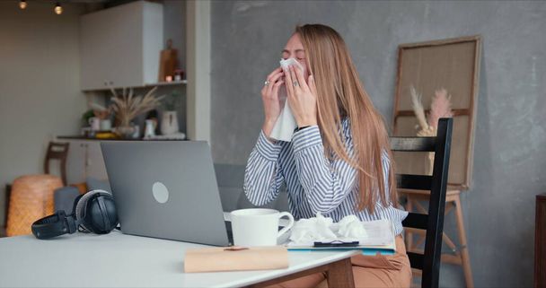 Sick young beautiful Caucasian woman coughs and sneezes with tissues, using laptop to work from home on quarantine. - 写真・画像