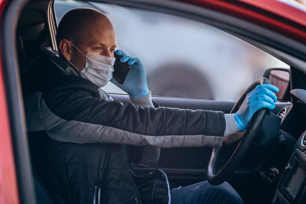 A man driving a car in protective medical mask and gloves is talking on phone. Safe drive in a taxi during a pandemic coronavirus. Protect driver and passengers from bacteria and virus infection. - Photo, image