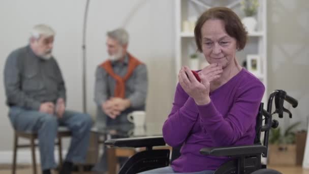 Positive elderly Caucasian woman looking at hand mirror and smiling as men chatting at the background. Pleasant female retiree getting ready to flirt with nursing home resident. - Filmagem, Vídeo