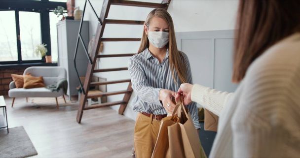 Young Caucasian woman wearing mask opens apartment door to get delivery bags from courier on self isolation lockdown. - Foto, Bild