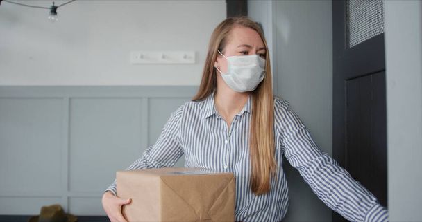 Young Caucasian woman in medical mask opens apartment door to get parcel delivery from courier during virus quarantine. - Foto, Bild