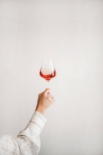 Womans hand in white shirt holding glass of rose wine over white wall background. Wine shop, wine tasting, bar, wine list concept - Foto, Imagen