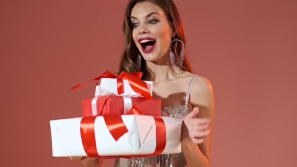 Happy pretty woman in bright sequins dress holding gift boxes and looking at the camera over red background - Кадры, видео