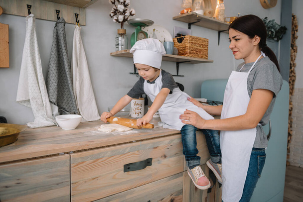 Молодая счастливая мама и ее ребенок готовят печенье дома на кухне. Рождественские пряничные пряники. милый мальчик с матерью в белой форме и шляпе приготовленные шоколадные печенья
 - Фото, изображение