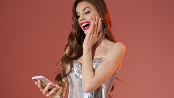 Kellemes csinos nő fényes flitteres ruha okostelefon használatával, és egyre boldogabb vörös háttér - Felvétel, videó