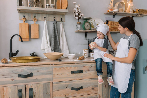 Young happy mom and her baby cook cookies at home in the kitchen. Christmas Homemade Gingerbread. cute boy with mother in white uniform and hat cooked chocolate cookies - Fotografie, Obrázek