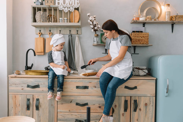 Young happy mom and her baby cook cookies at home in the kitchen. Christmas Homemade Gingerbread. cute boy with mother in white uniform and hat cooked chocolate cookies - Φωτογραφία, εικόνα