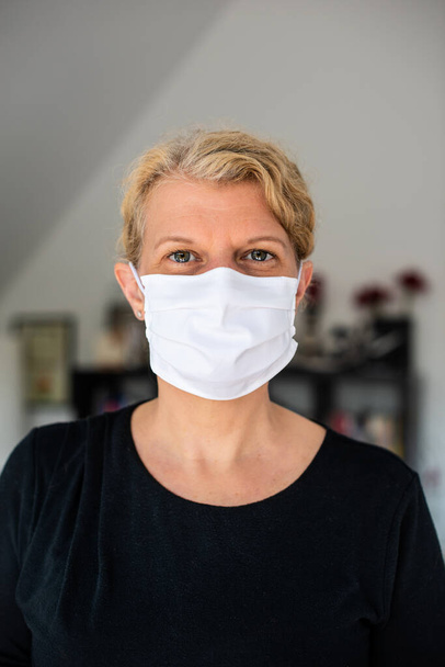 Πορτραίτο μιας μεσήλικας Καυκάσιας γυναίκας με μάσκα μέσης κατά του ιού της Κορόνας και λοίμωξη στο σαλόνι της στο σπίτι - Φωτογραφία, εικόνα