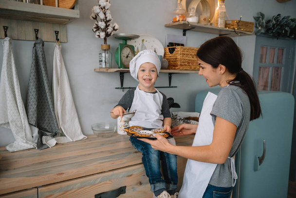 Young happy mom and her baby cook cookies at home in the kitchen. Christmas Homemade Gingerbread. cute boy with mother in white uniform and hat cooked chocolate cookies - Valokuva, kuva