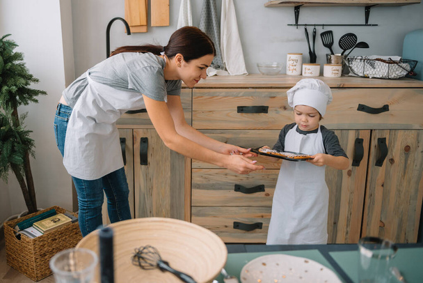 Young happy mom and her baby cook cookies at home in the kitchen. Christmas Homemade Gingerbread. cute boy with mother in white uniform and hat cooked chocolate cookies - Foto, imagen