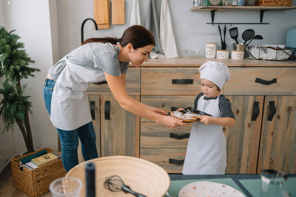 Young happy mom and her baby cook cookies at home in the kitchen. Christmas Homemade Gingerbread. cute boy with mother in white uniform and hat cooked chocolate cookies - Foto, Imagem