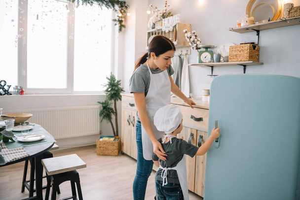 Young happy mom and her baby cook cookies at home in the kitchen. Christmas Homemade Gingerbread. cute boy with mother in white uniform and hat cooked chocolate cookies - Fotografie, Obrázek