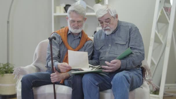 Hombre mayor caucásico mostrando viejas fotos en blanco y negro a un amigo en un asilo. Dos jubilados maduros positivos en anteojos compartiendo recuerdos en interiores
. - Imágenes, Vídeo