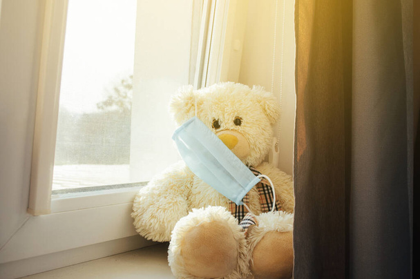 Coronavirus epidemic, COVID-19. The bear sits in a medical mask on the windowsill and looks at the camera. The concept of worldwide isolation is driven by a pandemic. Stay at home. - Zdjęcie, obraz