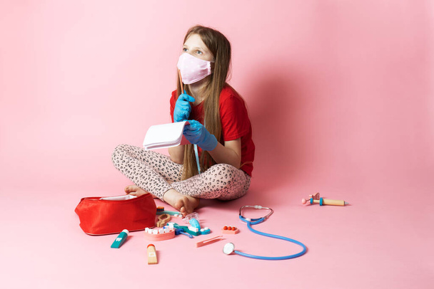 Personal protective equipment: a teenager girl in a medical mask and blue gloves plays a nurse and holds a stethoscope. Portrait on a pink background. - Photo, Image
