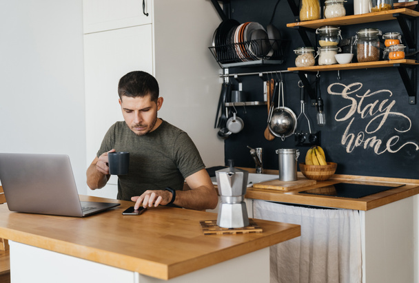 Atrakcyjny młody człowiek pracujący na laptopie we wnętrzu domowej kuchni i przytulnej atmosferze rano. Praca zdalna. - Zdjęcie, obraz