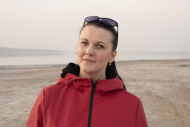 A brunette woman in sunglasses and a red jacket on an empty beach during quarantine. - Photo, Image