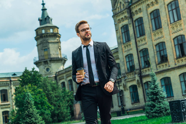 Handsome smiling young businessman wearing suit walking outdoors at the city streets, holding coffee cup - Fotografie, Obrázek