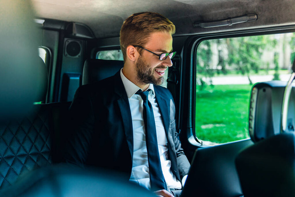 Handsome smiling young man in full suit and eyeglasses working using laptop while sitting in the car - Photo, Image