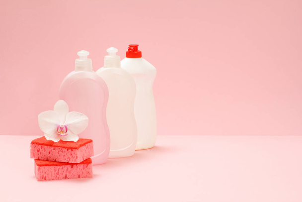 Пластиковые бутылки жидкости для мытья посуды, стеклоочиститель и моющее средство для микроволновых печей и печей, губки и белый цветок орхидеи на розовом фоне. Набор для стирки и очистки
. - Фото, изображение