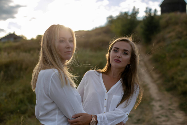 twee prachtige langharige volwassen zussen in witte blouses staan omarmd op een grazig heuvelpad op het platteland - Foto, afbeelding