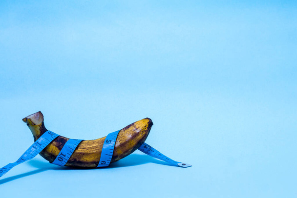Conceito de pênis velho sobre banana madura com fita de medição azul envolto em torno isolado em um fundo azul o tamanho do conceito importa
 - Foto, Imagem