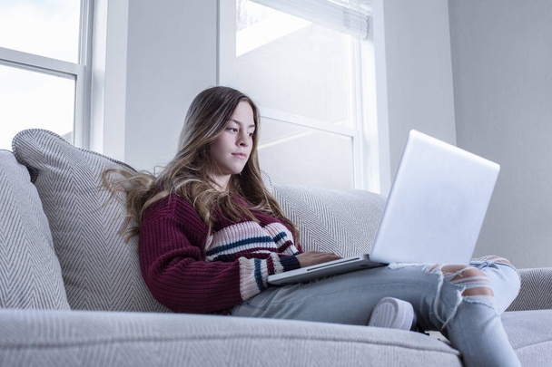 Έφηβη κοπέλα σπουδάζει στο σπίτι χρησιμοποιώντας φορητό υπολογιστή, κάθεται σε άνετο καναπέ. - Φωτογραφία, εικόνα