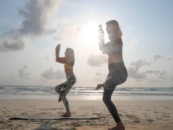 Kaksi naista harjoittelee kotka-joogaa meren rannalla auringonpaisteessa
 - Valokuva, kuva