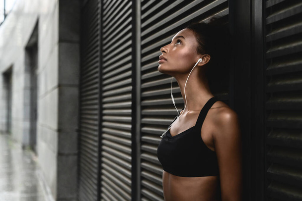 Vista lateral da mulher esportiva ouvindo música em fones de ouvido e inclinando-se para uma parede ao ar livre fazendo uma pausa após o treino
 - Foto, Imagem