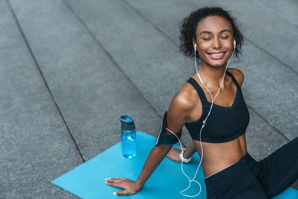 Horizontal shot of cheerful fitness woman relaxing after workout and listening music outdoors - Foto, imagen