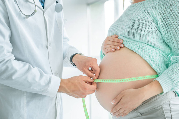 Side view of a pregnant woman middle body section exposing her belly while doctor uses a measuring tape to follow the growth of the baby at a hospital, indoors. - Photo, Image
