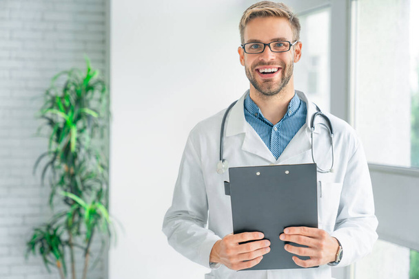 Handsome smiling medical doctor in white coat with clipboard, looking at camera and smiling while standing in clinic - Photo, image
