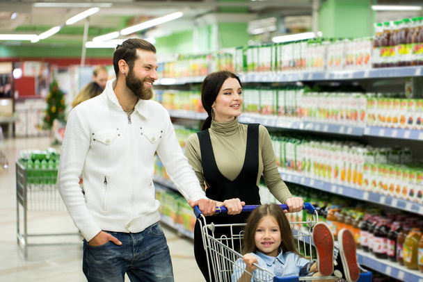 Krásné mladé rodiče a jejich roztomilé dcerušky se usmívají při výběru jídla v supermarketu. Otec tlačí nákupní vozík, zatímco dívky sedí tam. - Fotografie, Obrázek
